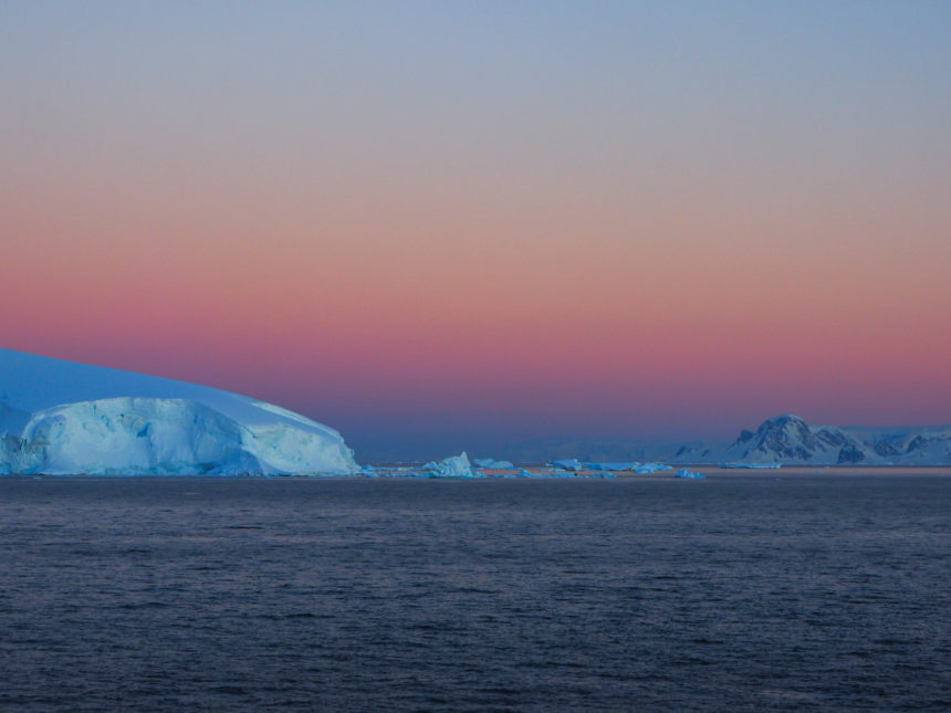 Über den Neumayer-Kanal und den Eisbergen färbt sich der Himmel bei Sonnenuntergang in ein rosa Licht.