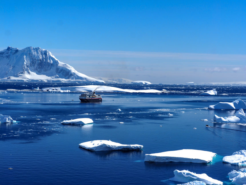 Bei Cuverville Island schippert bei strahlendem Sonnenschein ein Schiff vorbei an vereinzelten Eisschollen im Südpolarmeer.