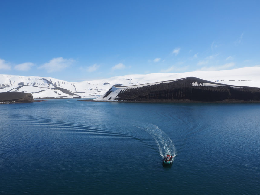 A tender boat cruises through the dark blue waters past snow-capped volcanoes at Deception Island, Antarctica.