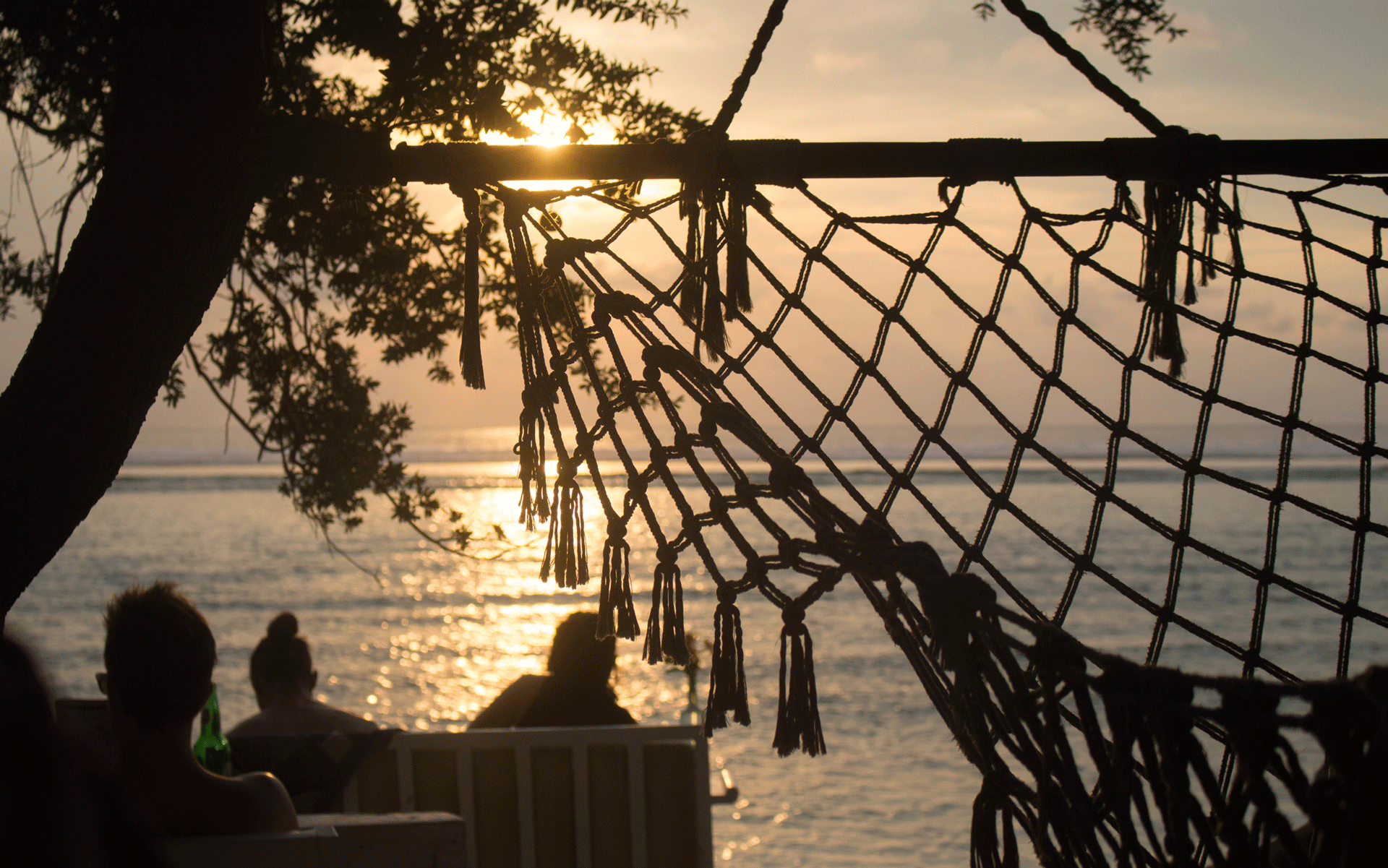 Vor einer gespannten Hängematte des Casa Vintage Beach Restaurants auf Gili Trawangan im Westen der Gili Inseln sitzen Menschen am Strand und beobachten den Sonnenuntergang.