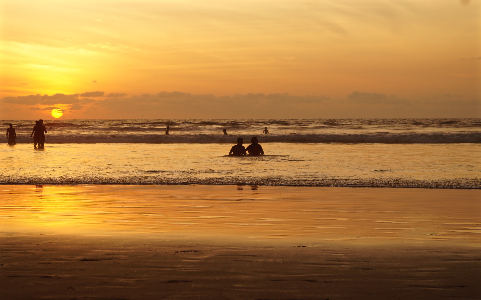 Der Sonnenuntergang am Horizont taucht den Strand in ein sanftes gelbes Licht, vereinzelt sind Menschen im Wasser.