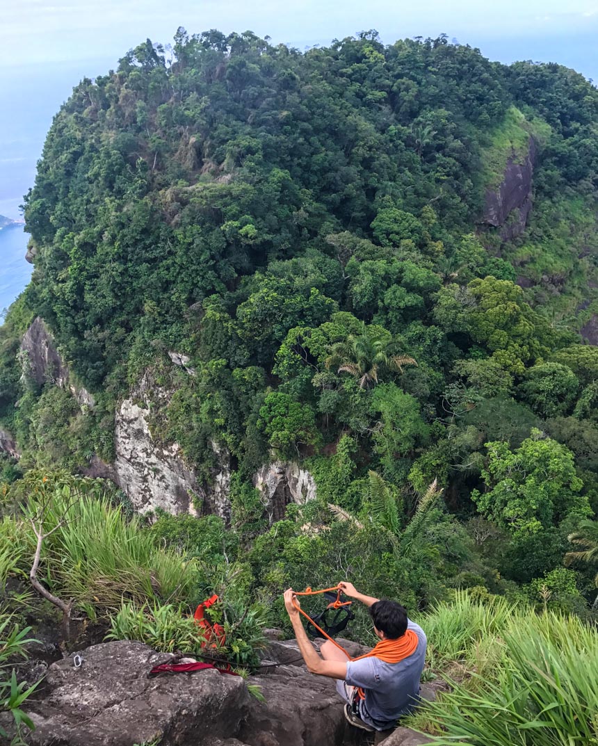 Ein brasilianischer Guide sichert bei Pedra da Gavea am Abgrund eines dichtbewachsenen Berges mit einem orangen Seil den Gurt zum Aufstieg.