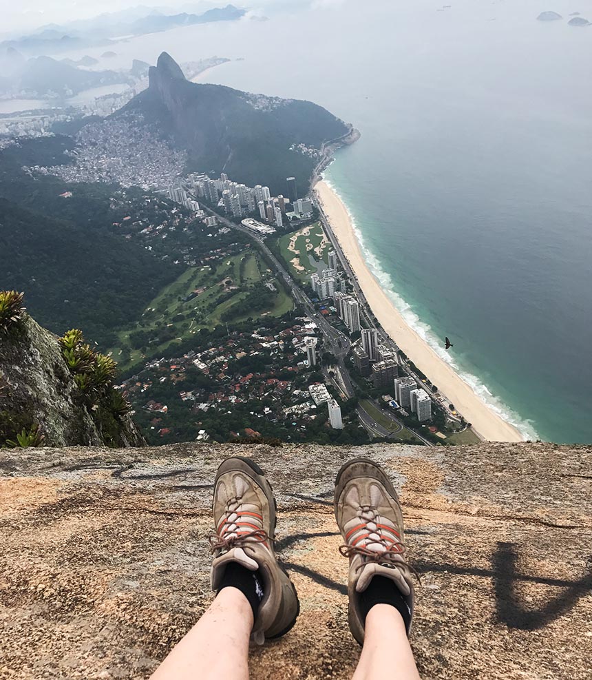 Zwei Beine mit Wanderschuhen baumeln über den Abgrund von Pedra da Gavea in Brasilien, unter ihnen eröffnet sich die Landschaft von Rio de Janeiro.