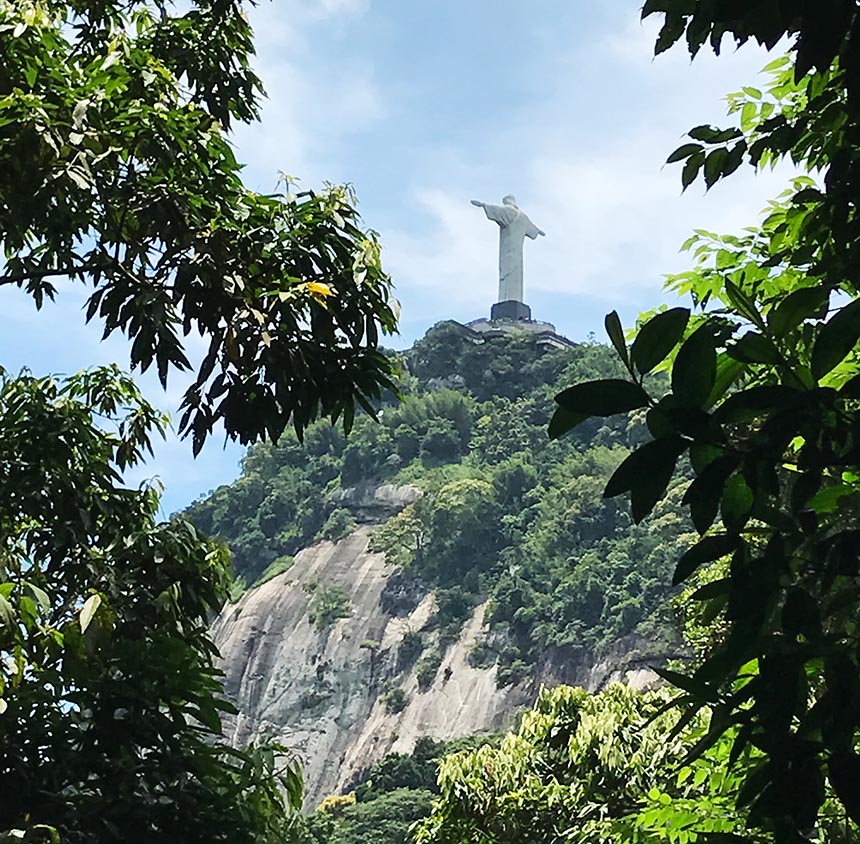 Zwischen Bäumen erblickt man die Christus Statue von hinten auf einem bewachsenen Berg.