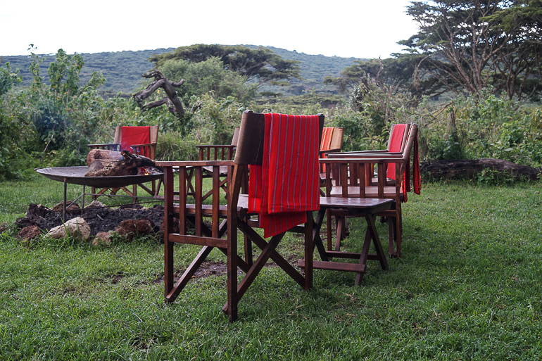 Auf einem Camp über dem Ngorongoro Krater sind drei Holzstühle mit roten Decken auf den Rückenlehnen um eine Feuerstelle postiert.