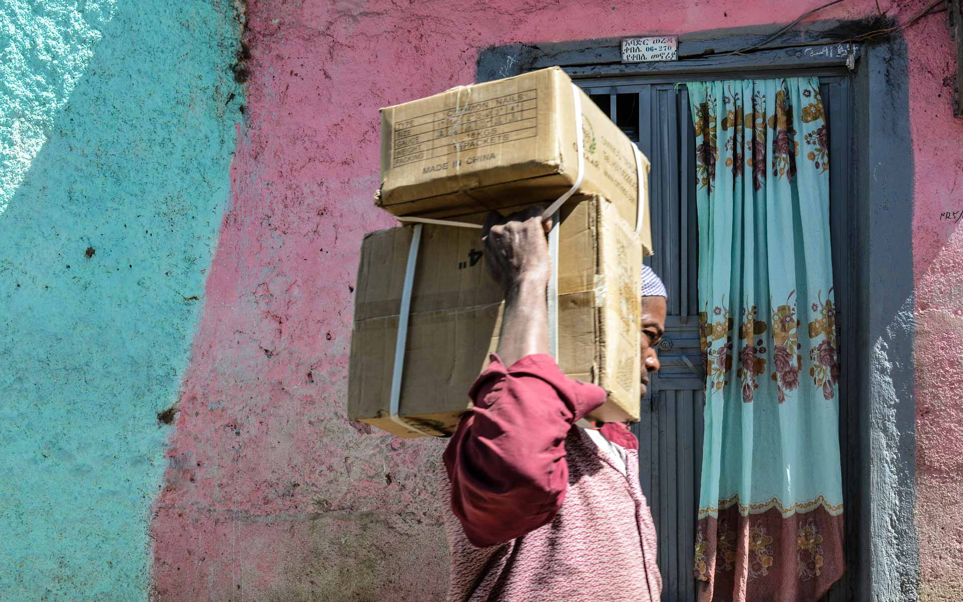 In Harar, Äthiopien, trägt ein dunkelhäutiger Mann vor einer blau-lila Häuserwand zwei Pakete auf der Schulter durch die Straße.