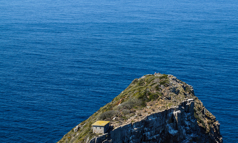 Am Kap der guten Hoffnung sieht man von oben einen Felsen, der aus dem Meer ragt und die Wassermassen des Indischen und des Atlantischen Ozeans, die hier in verschiedenem blau aufeinander treffen.