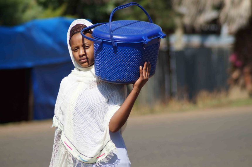Ein einheimisches Mädchen in weiß gekleidet, trägt auf ihrer linken Schulter einen großen blauen Topf entlang der Straßen von Kafa in Äthiopien.
