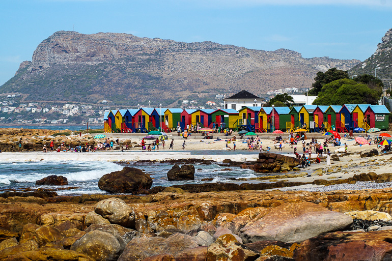 Bunte Strandhäuschen reihen sich an einem felsigen Strand bei Fishoek in Südafrika.