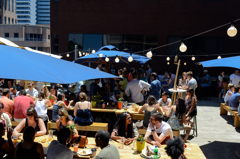 Unter blauen Sonnenschirmen des Neighbourgoods Market sitzen junge Menschen an Holztischen und essen, trinken und unterhalten sich.