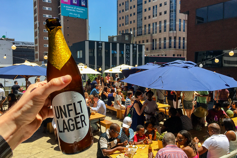 Auf dem Neighbourgoods Market hält der Fotograf ein Bier Richtung Sonnenterasse auf der Menschen unter blauen Sonnenschirmen sitzen.