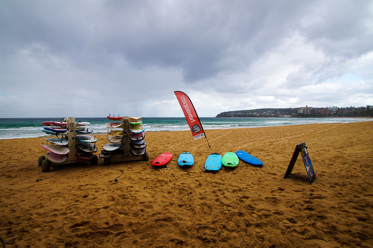 Am gelben Sandstrand von Manly, Australien, liegen Surfboards einer Surfschule trotz bewölktem Himmel für ihre Surfer bereit.
