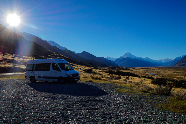 Ein weißer Camper-Van steht umgeben einer traumhaften Bergkulisse von Neuseeland im Licht der Sonne.