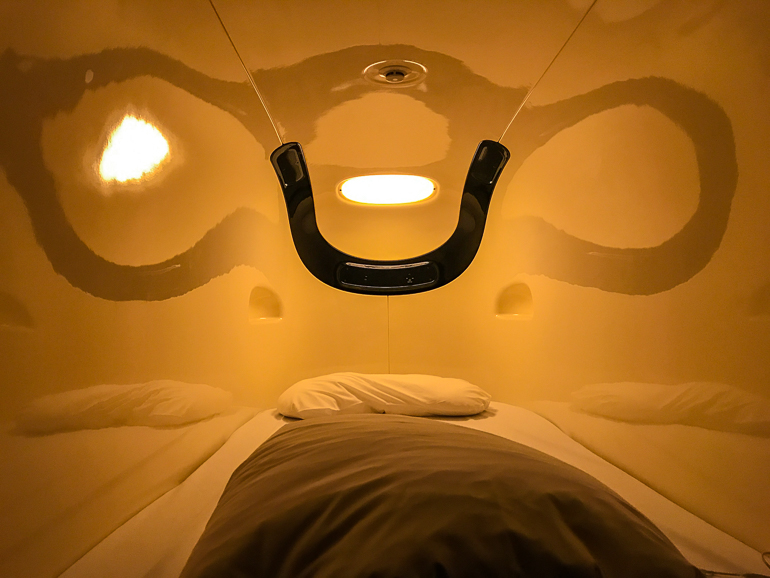 Das Innenleben des Kapselhotels in Tokio zeigt drei Schlafstätten mit je einem Kissen und einer Decke inmitten gelber beleuchteter Wände.