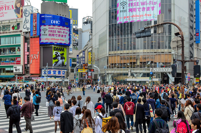 Inmitten der Kreuzung im Herzens Shibuya, Japan tummeln sich viele Menschen; die Häuserfassaden sind übersäht von allerlei Reklamen und Werbeschildern.