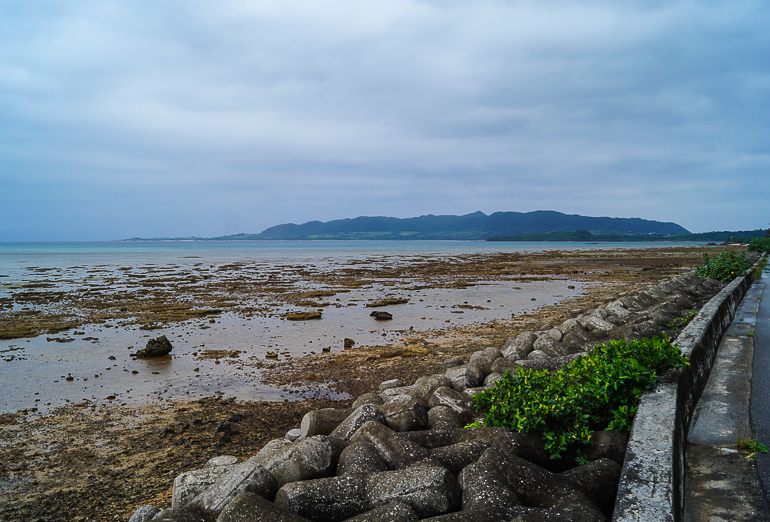 Auf der Yaeyama Insel Ishigaki in Japan trübt der Regen den Blick auf das mit Steinen durchzogene Meeresufer und den bewachsenen Hügeln am Horizont.