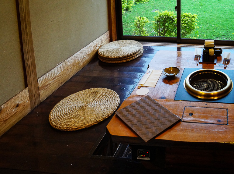 In einem kleinen Raum des Restaurants Ishigakiya auf der Yaeyama Insel Ishigaki in Japan wird den Gästen auf einem dunklen Holzboden mit Bambussitzkissen und bodentiefen Fenstern mit Blick zum Garten landestypische Rindfleischspezialitäten serviert.