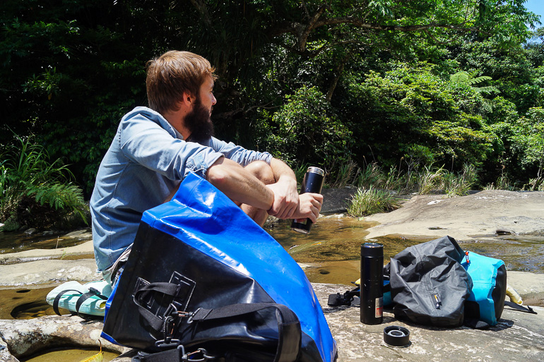 Der Blogger Clemens sitzt in der Nähe der Sangara Wasserfälle in Iriomote, Japan zwischen Rucksäcken auf Steinen im Wasser und hält eine geöffnete Thermoskanne in der Hand.
