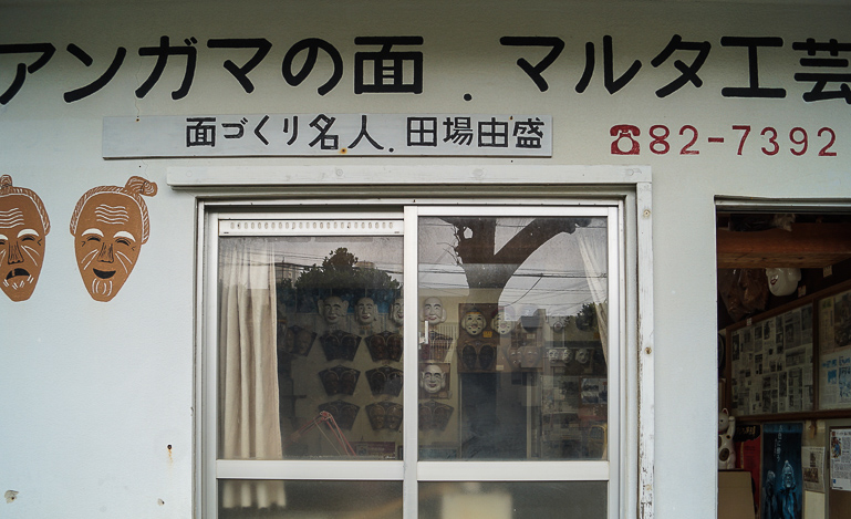 Die kleine Hausfassade mit einem Doppelfenster und geöffneter Tür, sowie blauer japanischer Schrift deutet auf das Büro des Maskenherstellers Mr. Taba auf der Yaeyama Insel Ishigaki in Japan hin.