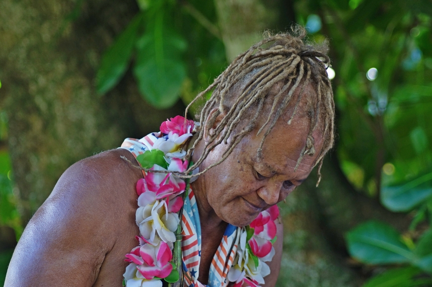 Rarotongas berühmtester Medizinmann Pa hat unter Palmen seinen Blick zum Boden gerichtet, um seinen Hals trägt er eine Blumenkette und ein Halstuch.