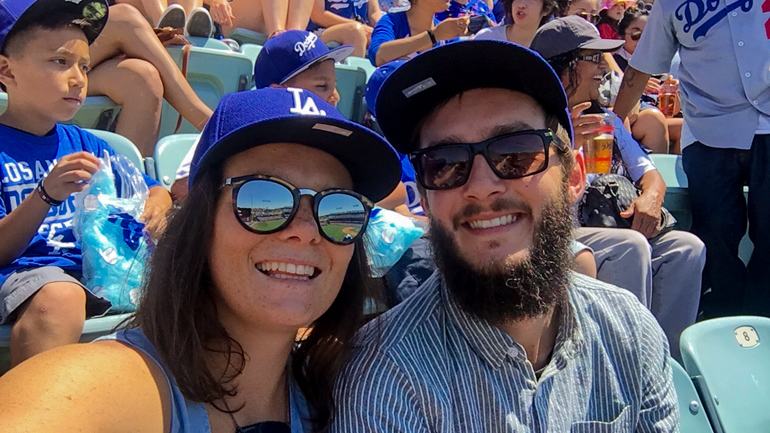 Freudestrahlend blickt ein Pärchen während eines L.A. Dodgers Baseballspiels in die Kamera, beide tragen blaue L.A.-Caps.