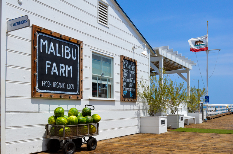 Das Malibu Farm Restaurant mit seiner weiß getünchten Fassade befindet sich am Malibu Pier, ein kleines Wägelchen an der Hauswand ist gefüllt mit Wassermelonen und eine Los Angeles Fahne weht im Wind.