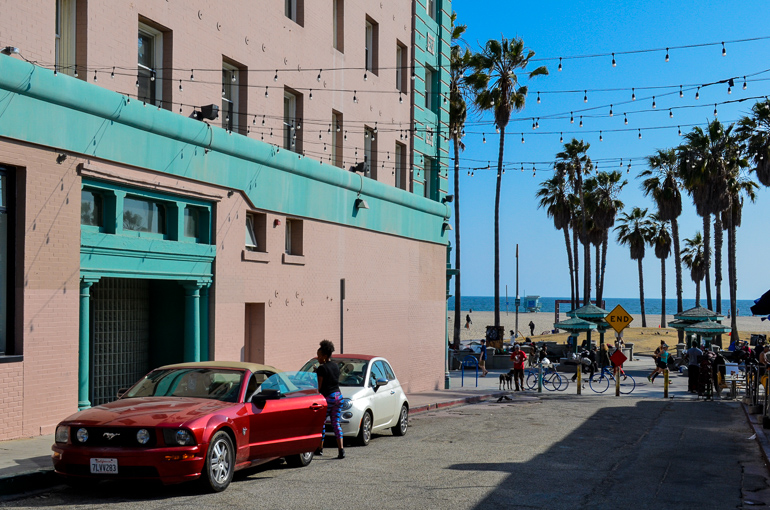 Eine Seitenstraße in Santa Monica am Venice Beach zeigt eine rosa-grüne Hausfassade, eine Frau steigt gerade in ihren roten Ford Mustang, Lichterketten und Palmen ragen in den Himmel.