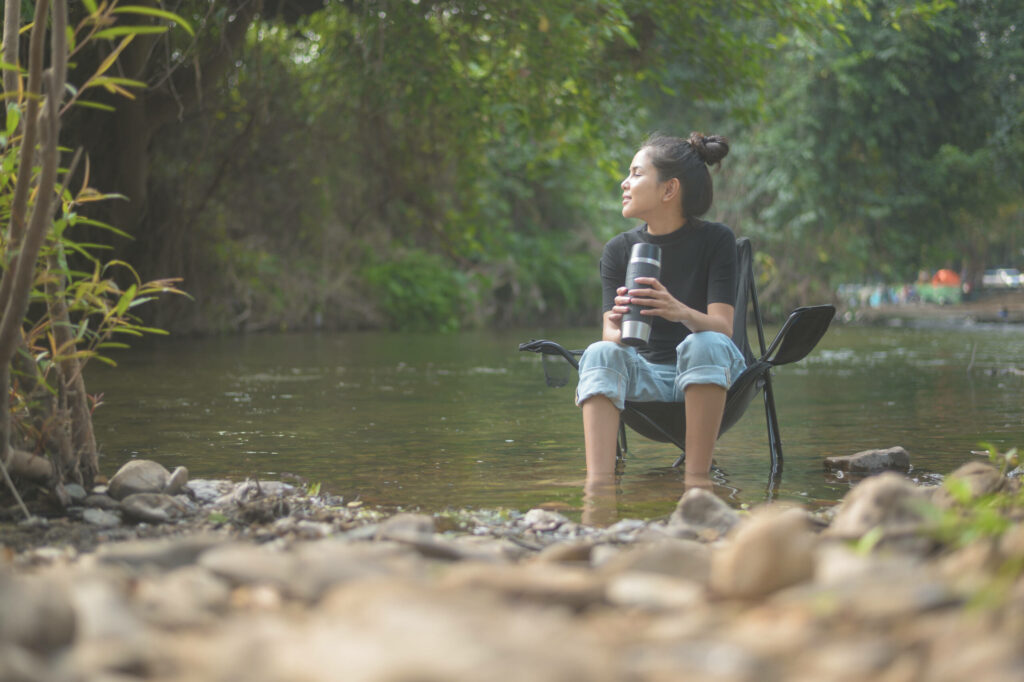 Idyllische Umgebung am See, Frau entspannt im Campingsitz am Wasser mit einem nützlichen Kaffee-Gadget.