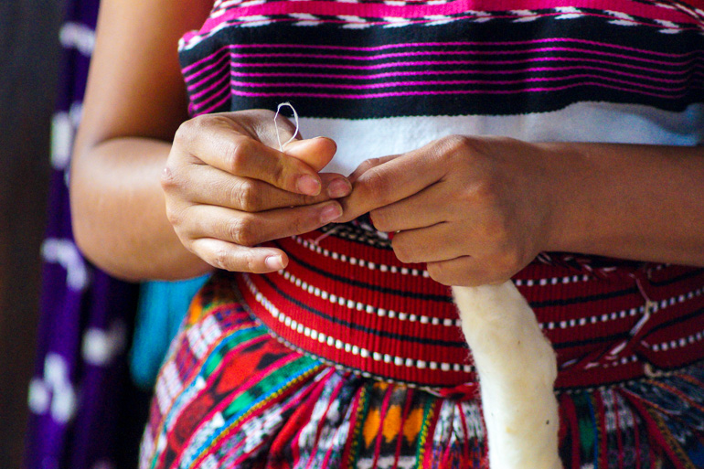 Eine bunt gekleidete, einheimische Frau verarbeitet mit ihren beiden Händen Baumwolle an einer länglichen Schnur.