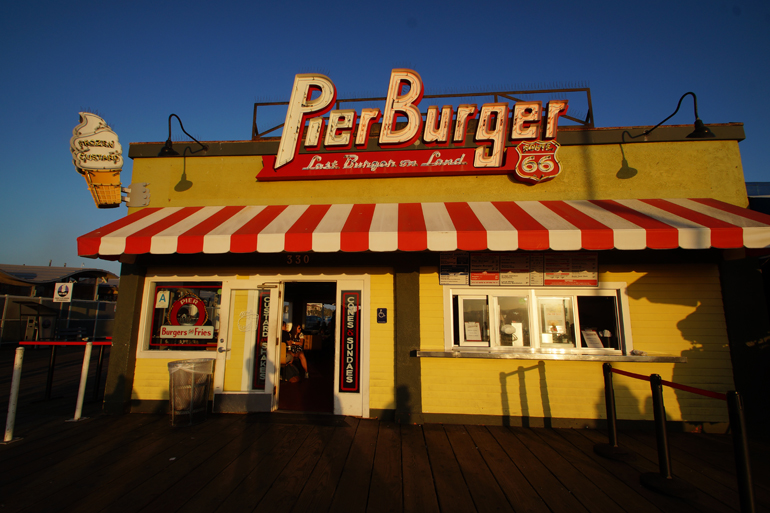 Ein gelbes Burger Haus mit der Aufschrift Pier Burger und rot-weiß gestreifter Markise bei Sonnenuntergang in Santa Monica.