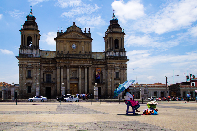 In Guatemala Stadt sitzt am Central-Plaza, vor der Kulisse einer Kirche, eine Snackverkäuferin unter einem blau-karierten Sonnenschirm.
