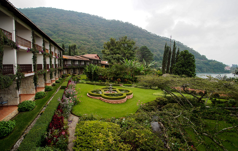 Das Hotel Atilan in Panajachel besitzt einen parkähnlichen Hotelgarten, im Hintergund strotzt unter bewölktem Himmel ein bewachsener Berg.
