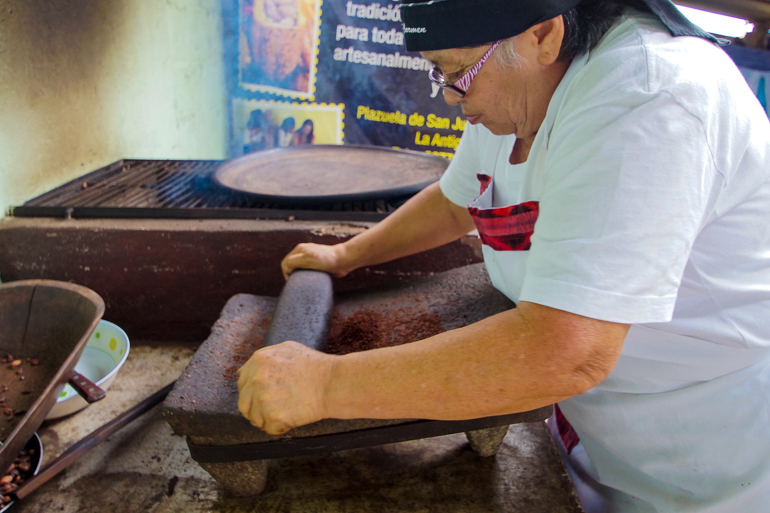 Eine einheimische Dame in Guatemala mahlt auf einem steinernen Brett Schokoladenbohnen.