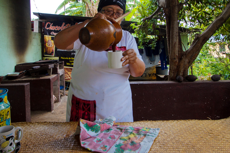 In einer Gartenküche gießt eine einheimische Frau aus einem braunen Tonkrug Schokolade in eine Tasse.