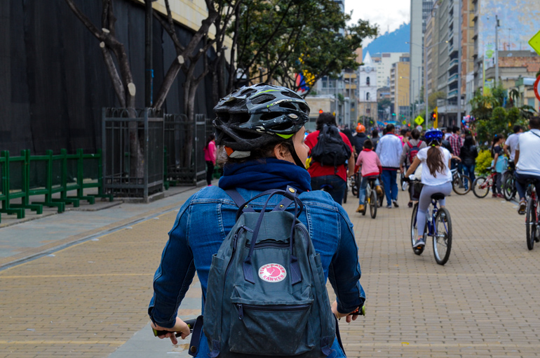 Auf einer Straße in Bogotá sind Fahrradfahrer unterwegs, eine Fahrradfahrerin mit Rucksack und in blau gekleidet blickt auf die Seite.