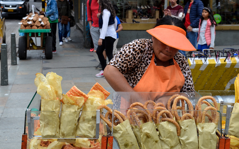 On Bogotá's streets, a local woman dressed in an orange apron and cap offers fried pastries for sale.