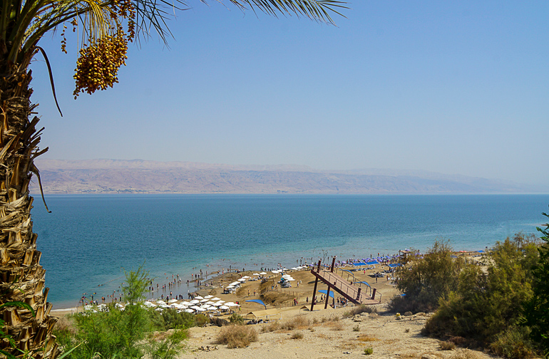 Am sandigen Ufer des Toten Meeres Kalia Beach in Israel tummeln sich unter weißen Sonnenschirmen und am Wasserrand Badegäste.