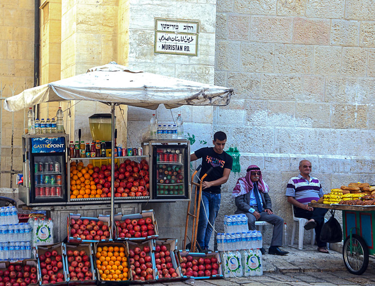An einem Häusereck in Jerusalem, Israel und unter einem weißen Sonnenschirm hat ein Händler einen Straßenstand mit Obst in Kisten zum Verkauf aufgebaut, zwei ältere Herren auf Plastikstühlen warten auf Kundschaft.