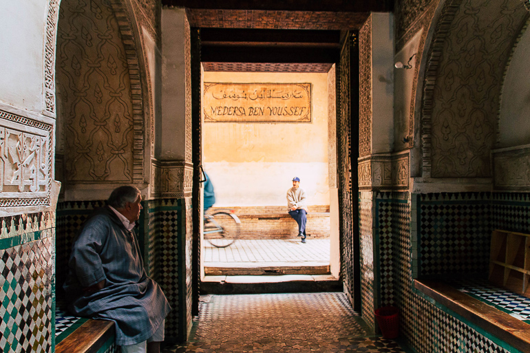 Der Eingangsbereich der alten Koranschule Ben Youssef in Marokko ist mit kleinen bunten Fliesen und Bemalungen verziert, ein älterer Herr hat sich zum Verweilen auf eine Bank unter einem Torbogen gesetzt, sein Blick ist auf die Straße gerichtet, auf der gerade ein Fahrradfahrer vorbeifährt.