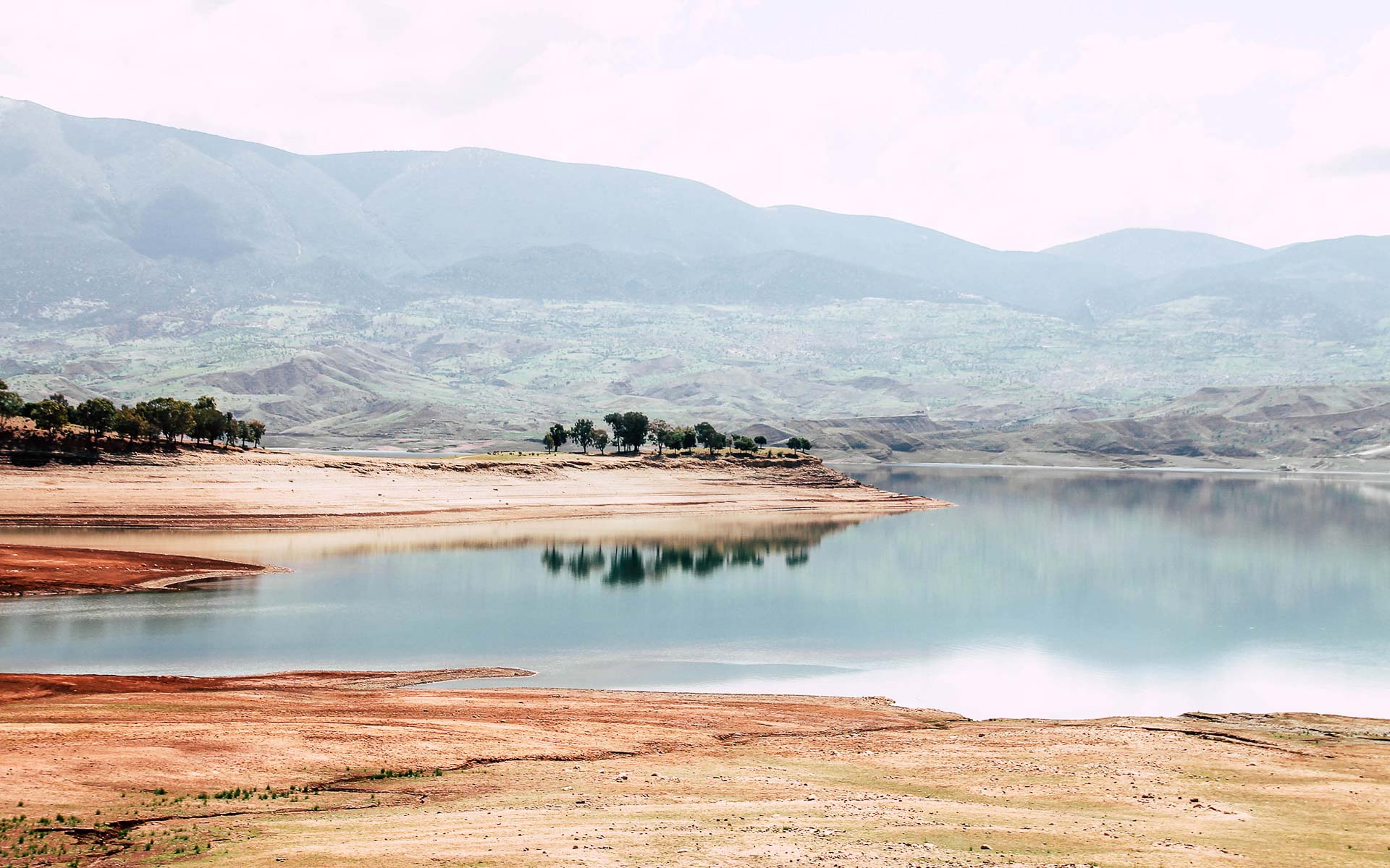 Das Ufer von Marokkos Staudamm Bin El Ouidane leuchtet in verschiedenen Rottönen, das Wasser spiegelt sich ruhig und klar in der Sonne, im Hintergrund liegen im Dunst grünbewachsene Hügel.