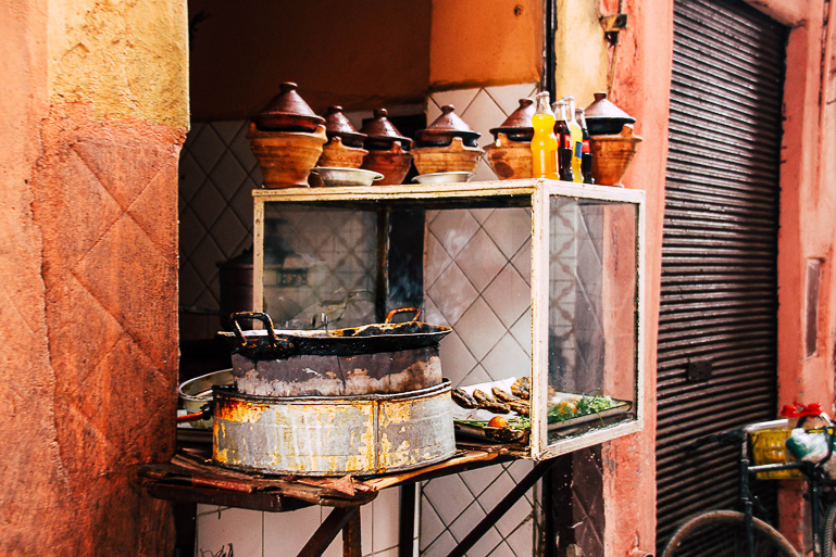 Inmitten einer roten Hauswand befindet sich in Marrakesch eine kleine Garkküche, der Kochtopf ist alt und eingebrannt, über einem Glaskasten wurden kleine Tontöpfchen mit spitzförmigen Deckeln angerichtet.