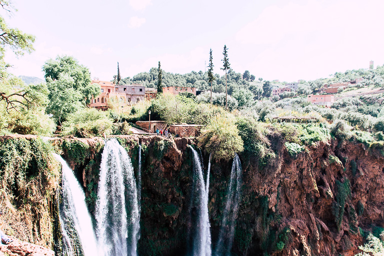 Hinter einem kleinen Dorf fließt an einem grün bewachsenen Berg in schmalen Rinnen das Wasser des Ouzoud Wasserfalls in Marokko hinab.