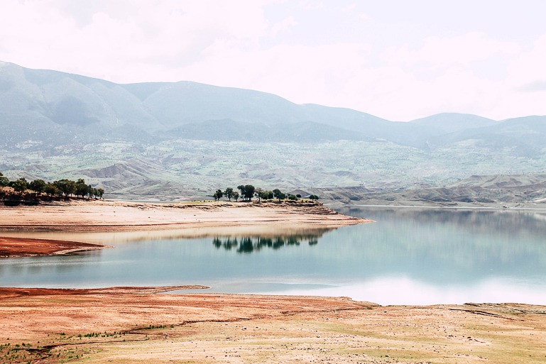 Das Ufer von Marokkos Staudamm Bin El Ouidane leuchtet in verschiedenen Rottönen, das Wasser spiegelt sich ruhig und klar in der Sonne, im Hintergrund liegen im Dunst grünbewachsene Hügel.