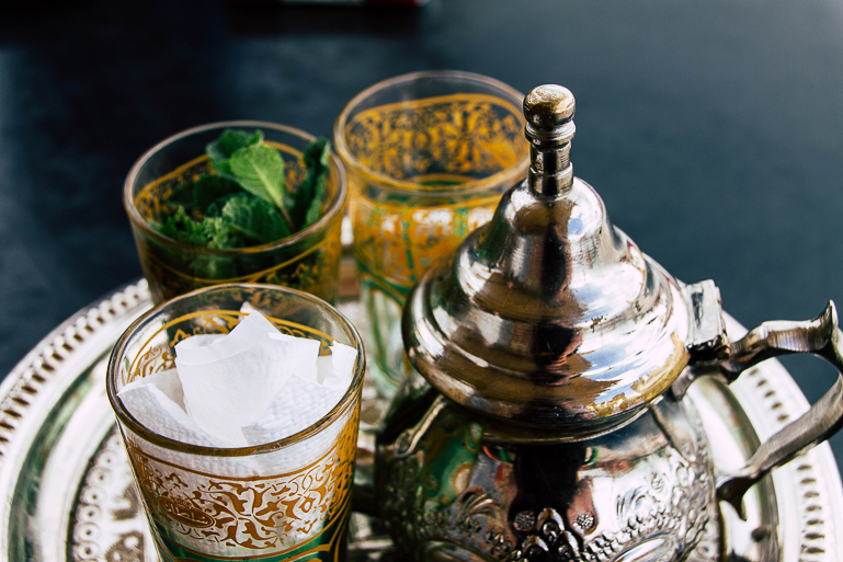 An antique teapot and three tea glasses are arranged on a silver tray, the tea glasses are filled with fresh mint for a typical Moroccan mint tea.