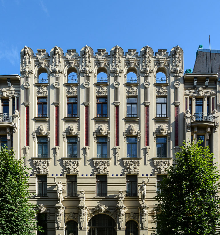 Bei der Fassade eines Gebäudes im perpendikularen Jugendstil in Riga, Lettland, wurden senkrechte Elemente hervorgehoben, ausgeprägte Erkerfenster und Reliefs sind in strengen Formen angeordnet und die Flächen zwischen den Stockwerken wurde mit Ornamenten verziert.