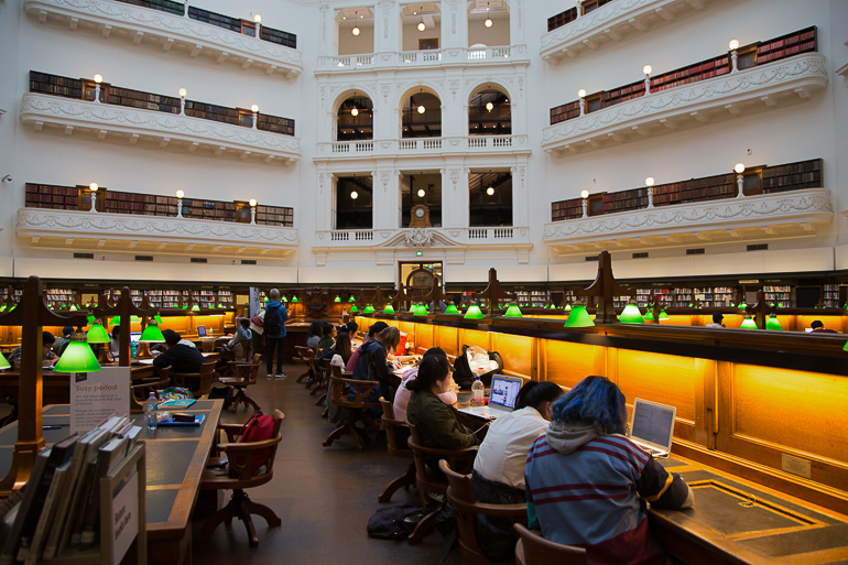 Viele Besucher sitzen an Schreibtischen mit grünen Leselampen inmitten der Public Library in Vicoria, Australiens Melbourne, die weißen Etagen sind stilvoll beleuchtet.