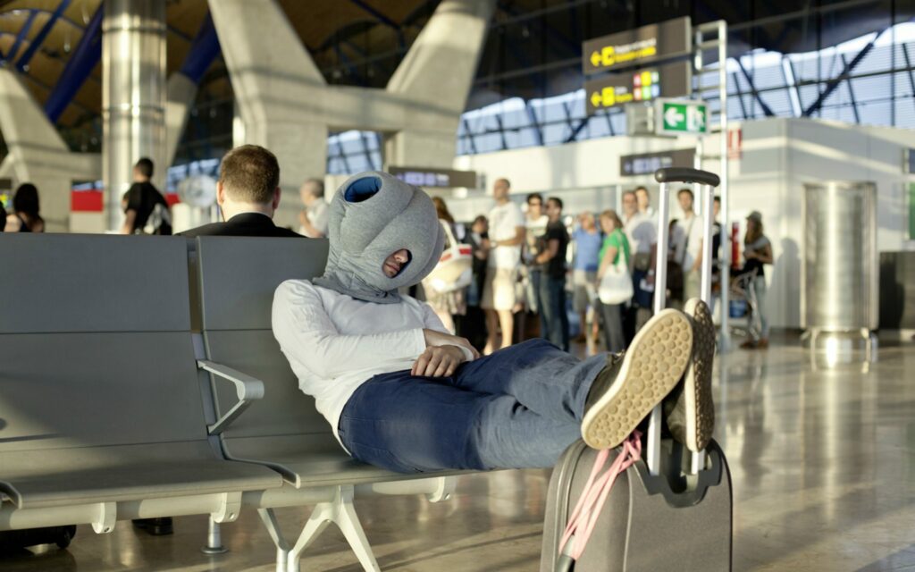 Ein Mann sitzt schlafend auf einer Stuhlreihe im Wartebereich eines Flughafens über seinem Kopf trägt er ein Ostrichpillow.