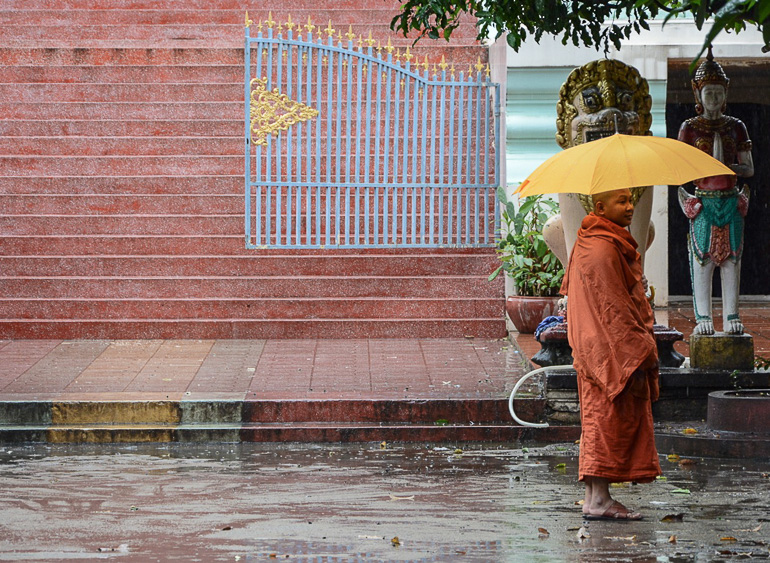 Ein Mönch in oranger Kleidung und Regenschirm spaziert barfuß auf der regennassen Straßen vor den Mauern des Lotus Tempels in Kambodscha, Phnom Penh.