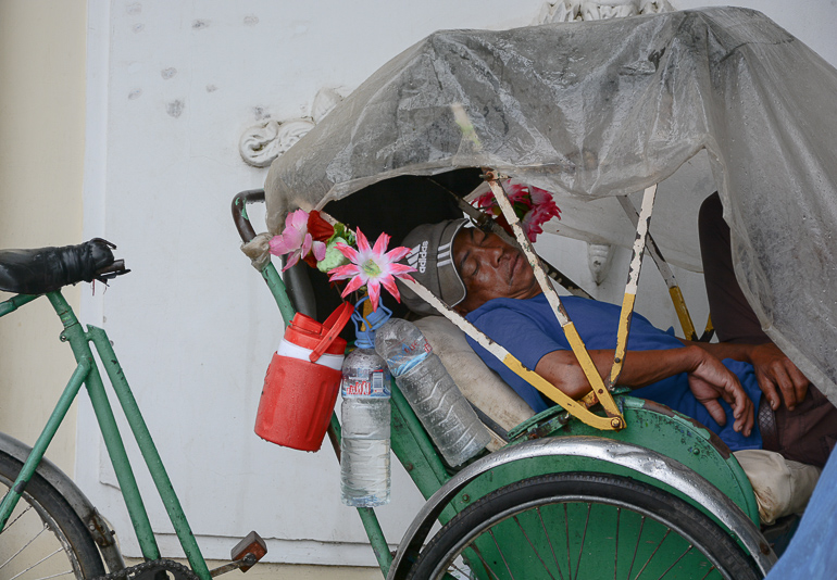 Ein Cyclo-Fahrer ruht an einer Hausmauer von Phnom Penhs Straßen in seiner grünen Rikscha, am Lenker sind eine Getränke und Blümchen befestigt.