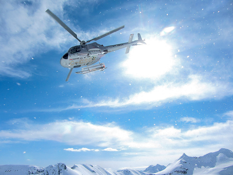 Ein Hubschrauber für Heli-Skiing kreist bei Sonnenschein über die verschneite Berglandschaft.